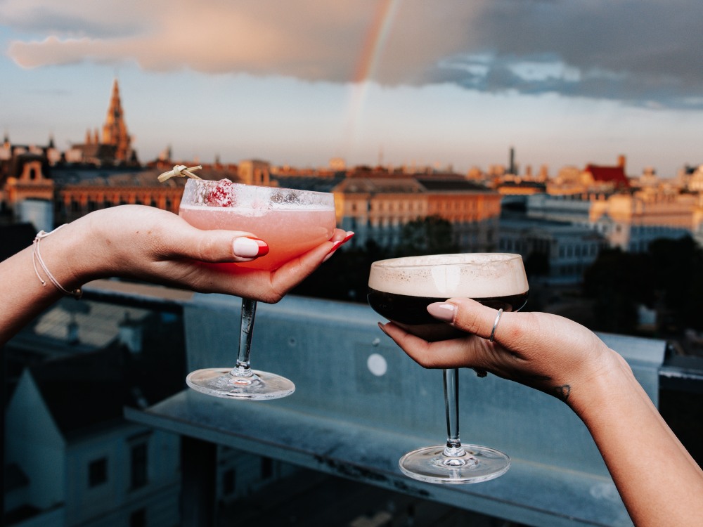 Two hands holding cocktails against a vibrant sunset sky with a rainbow, showcasing a city skyline in the background.