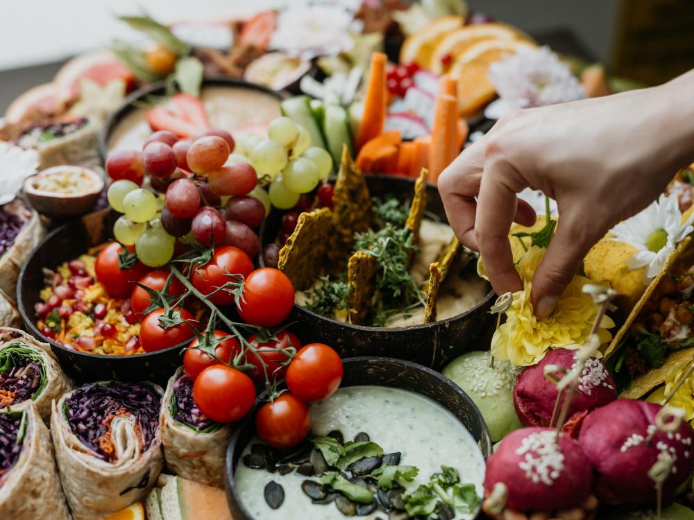 Eine gelbe Blume wird von Hand auf einer bunten Platte mit frischem Gemüse, Obst, Wraps, Dips und Crackern arrangiert.