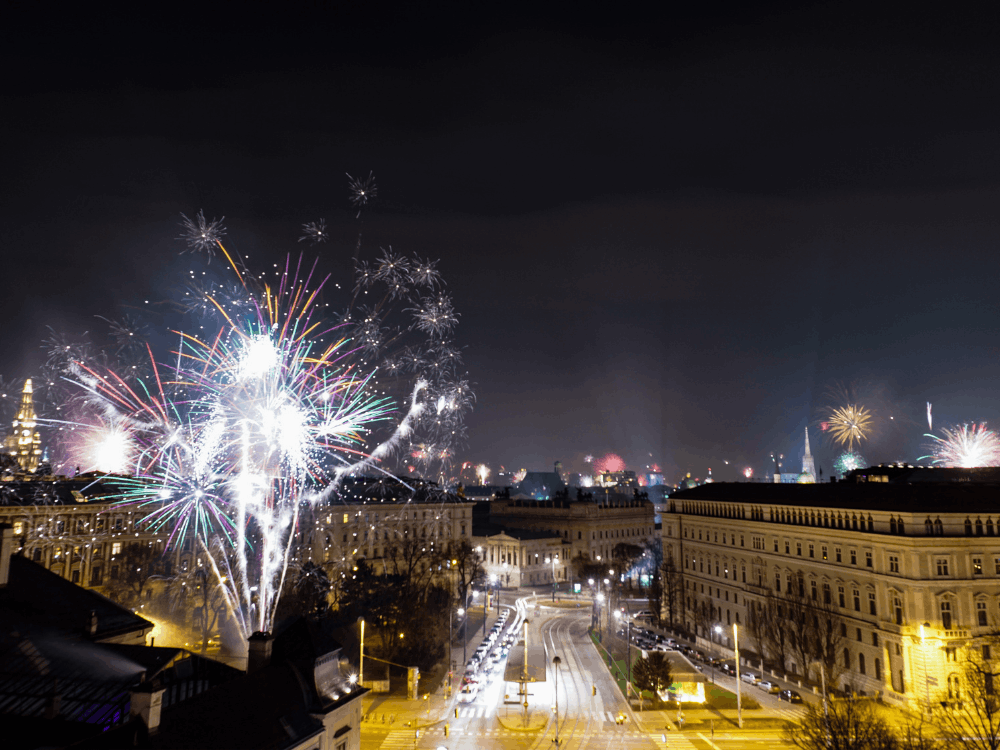 Spektakuläres, farbenfrohes Feuerwerk am Nachthimmel über historischen Gebäuden und einer beleuchteten Stadtstraße bei einer festlichen Feier.