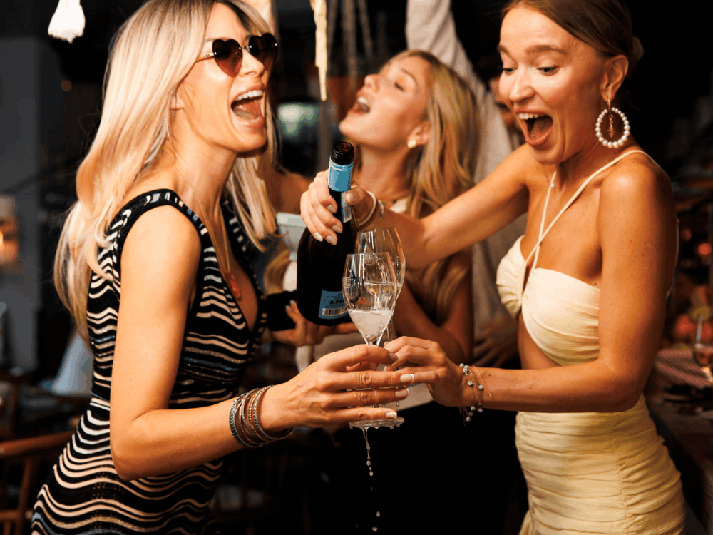 Drei ausgelassen feiernde Frauen lachen und stoßen mit Champagner an, während eine Flasche Schaumwein direkt in die Gläser eingeschenkt wird.